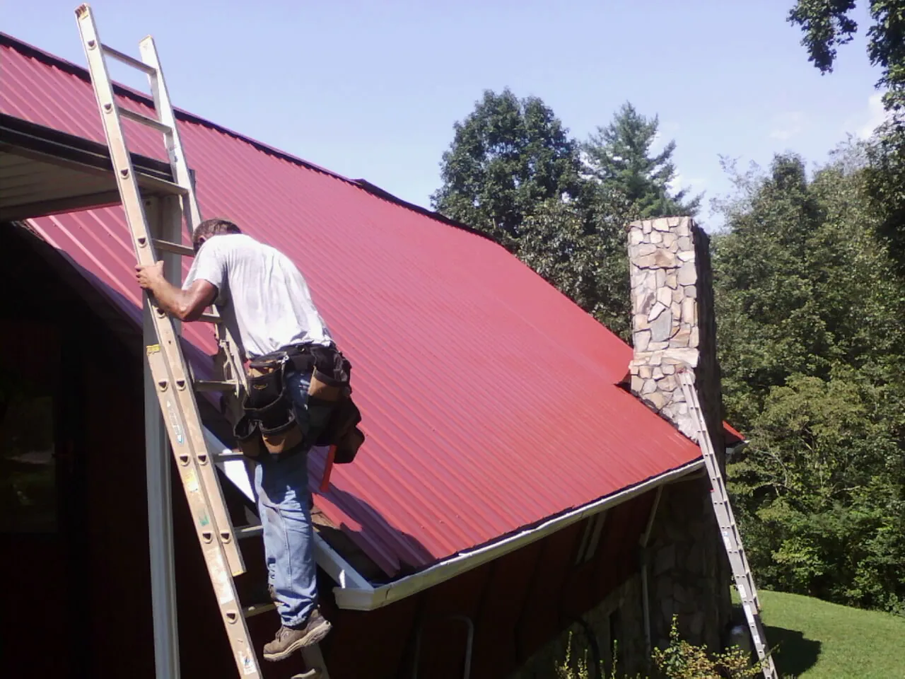 Roofer on a ladder beside a red metal roof and stone chimney — Soffit & Fascia Repair by Roofing Brothers in Whitestown